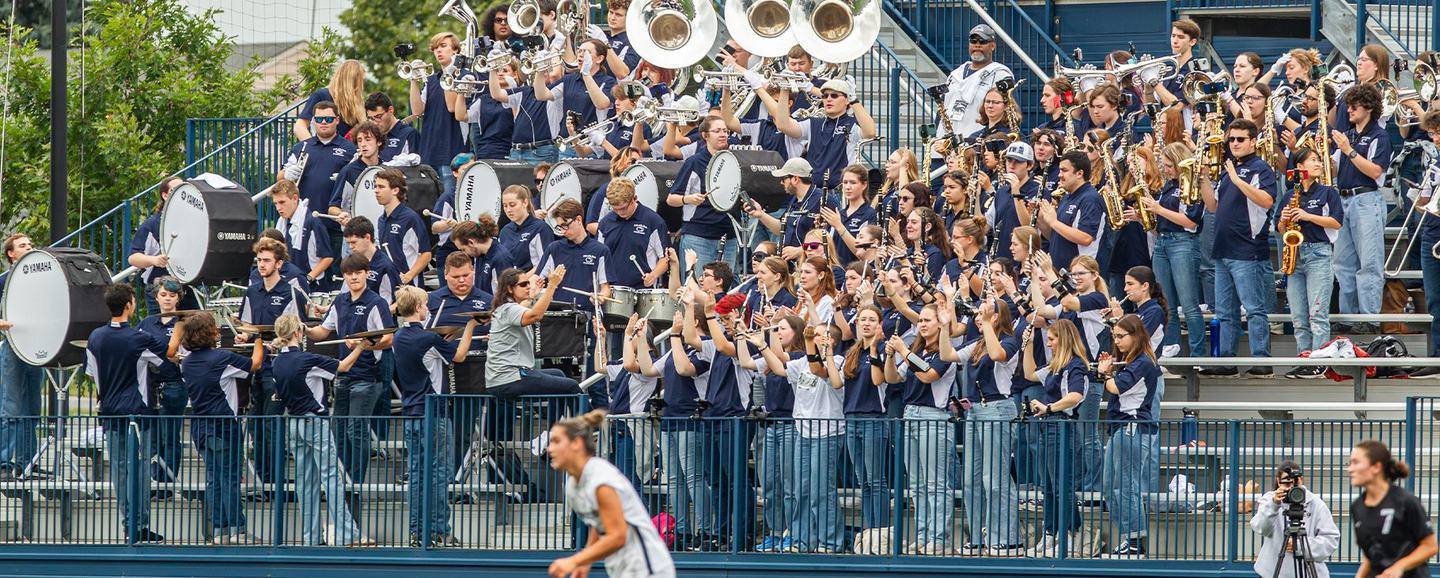 The Penn State Blue Band packed into the bleachers in daylight, horns up and bass drums along the front row, with players moving past below