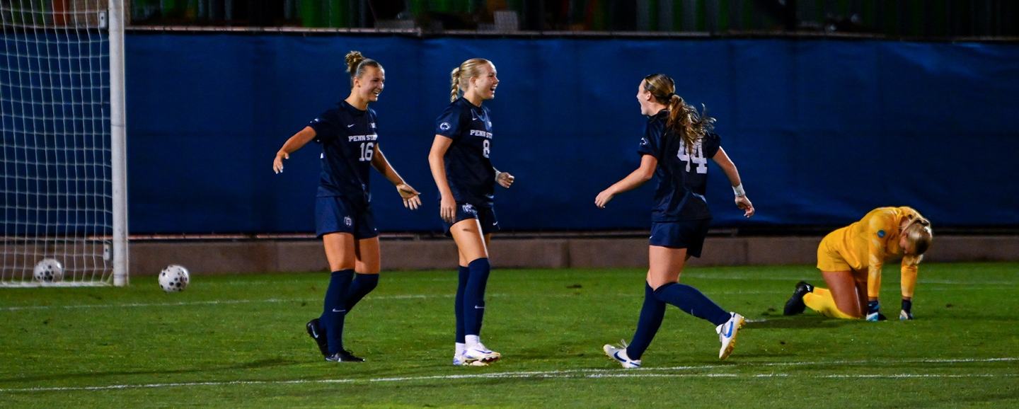 Three Penn State players in navy celebrate at night as the opposing goalkeeper kneels in a gold kit