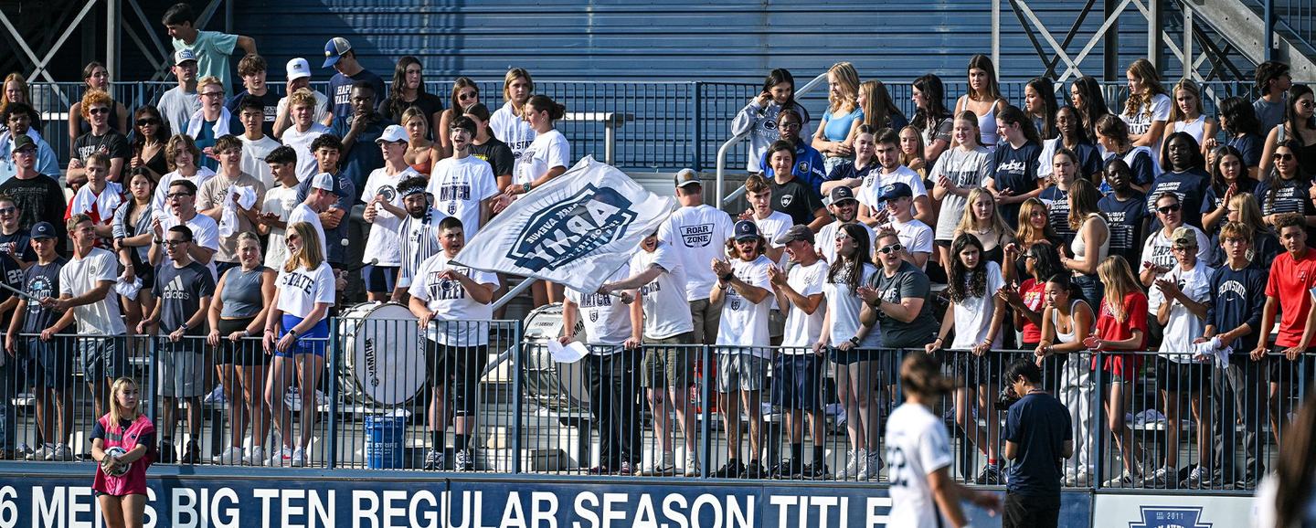 A packed student section in white t-shirts standing behind the blue touchline railings, one supporter waving a large Penn State flag